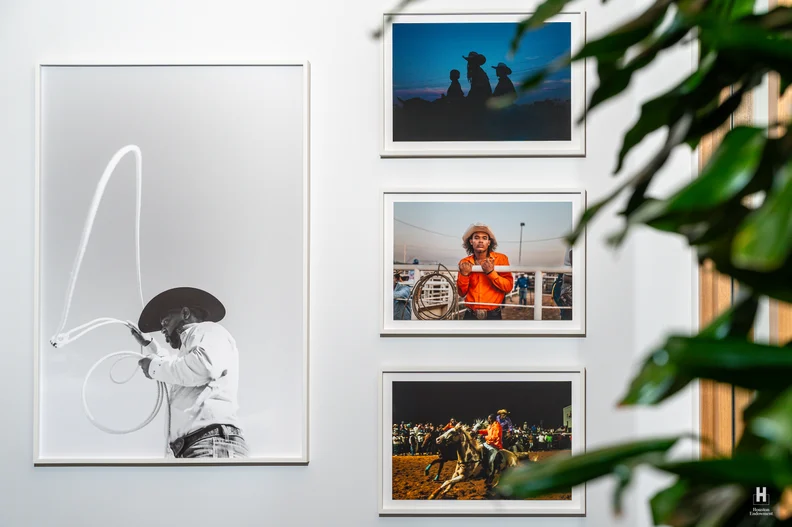 Framed photos on a white wall: large B&W cowboy swinging a lariat left, three color rodeo shots to the right, leafy plant partially covering.