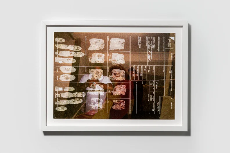 Framed sepia fingerprint card mounted on a gray wall, showing rows of white fingerprint impressions, labeled boxes and a faint reflection.